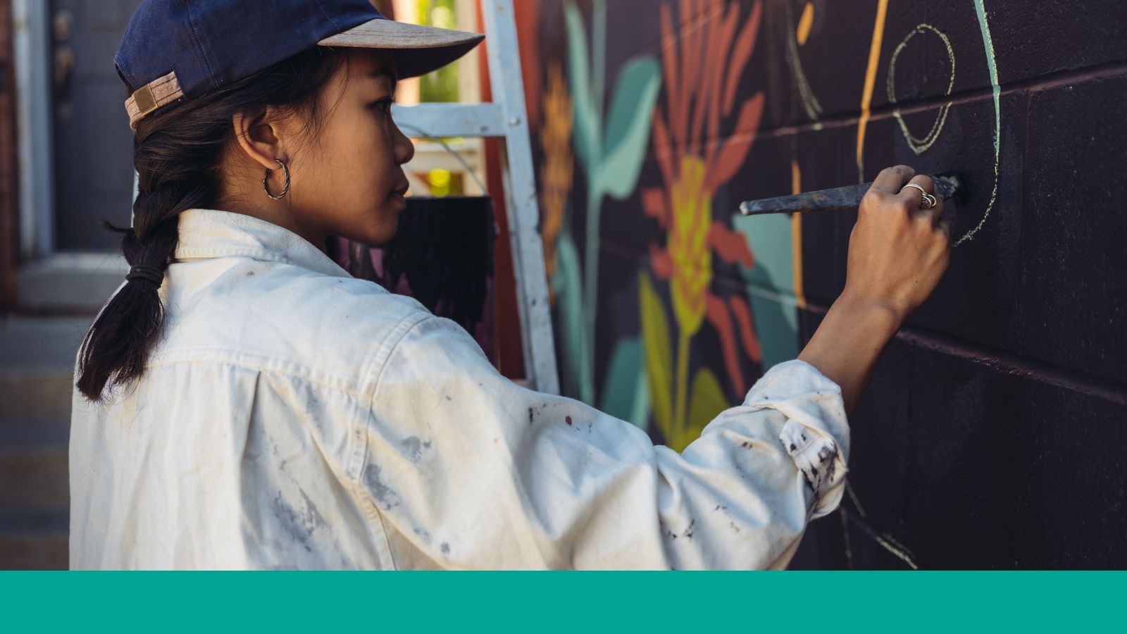 A woman in a baseball hat paints a mural.