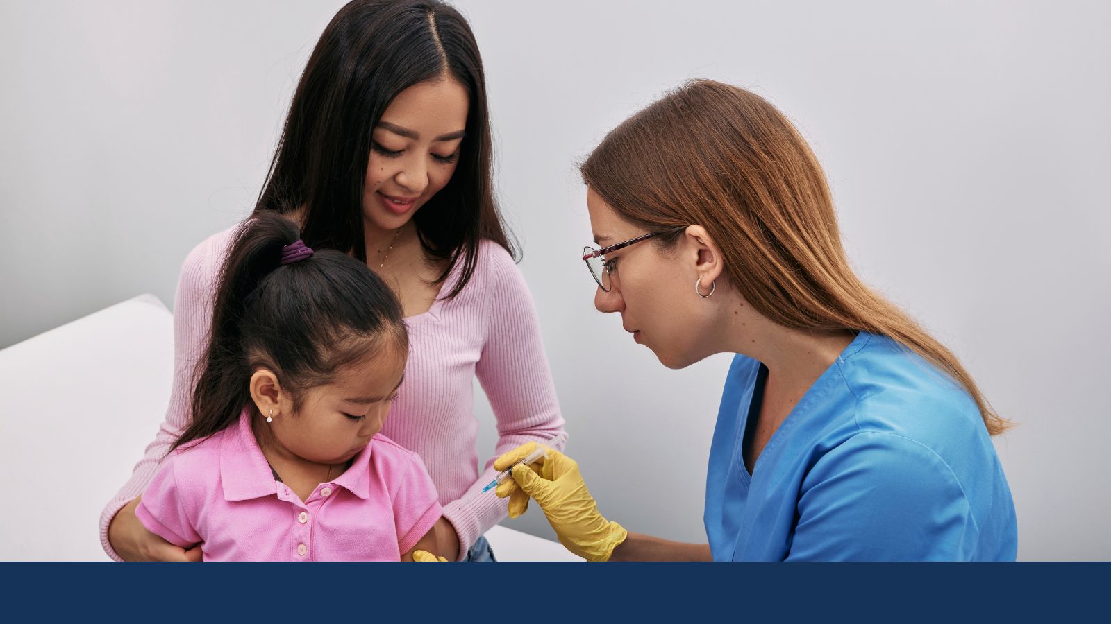 A medical professional gives a vaccine to a young child sitting on her caretaker's lap.